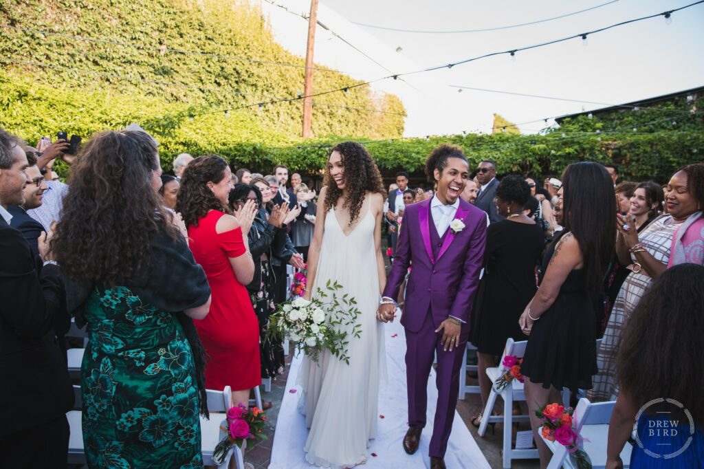 Bride and groom walking down the aisle laughing with guests cheering. Outdoor wedding ceremony at Seventh Place. Los Angeles wedding photojournalism by Drew Bird.