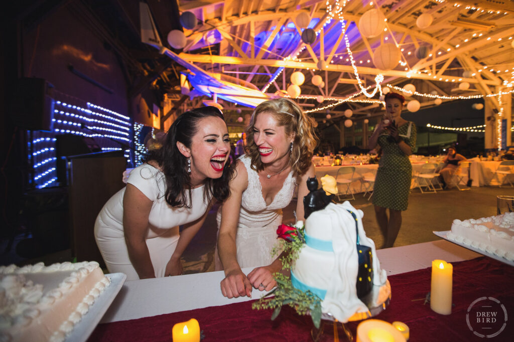 Two brides laughing at seeing wedding cake topper. Lesbian wedding. Gay wedding. Same-sex wedding. Oakland Aviation Museum wedding photographer Drew Bird.