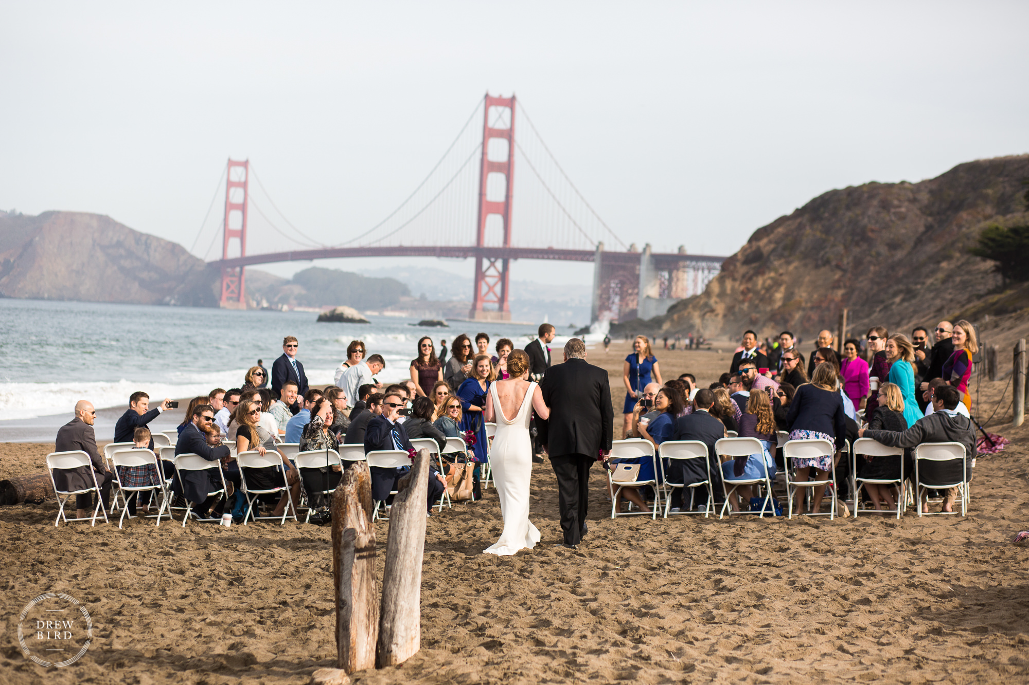 Bride and father walking down aisle. Baker Beach wedding ceremony in San Francisco with Golden Gate Bridge in background.