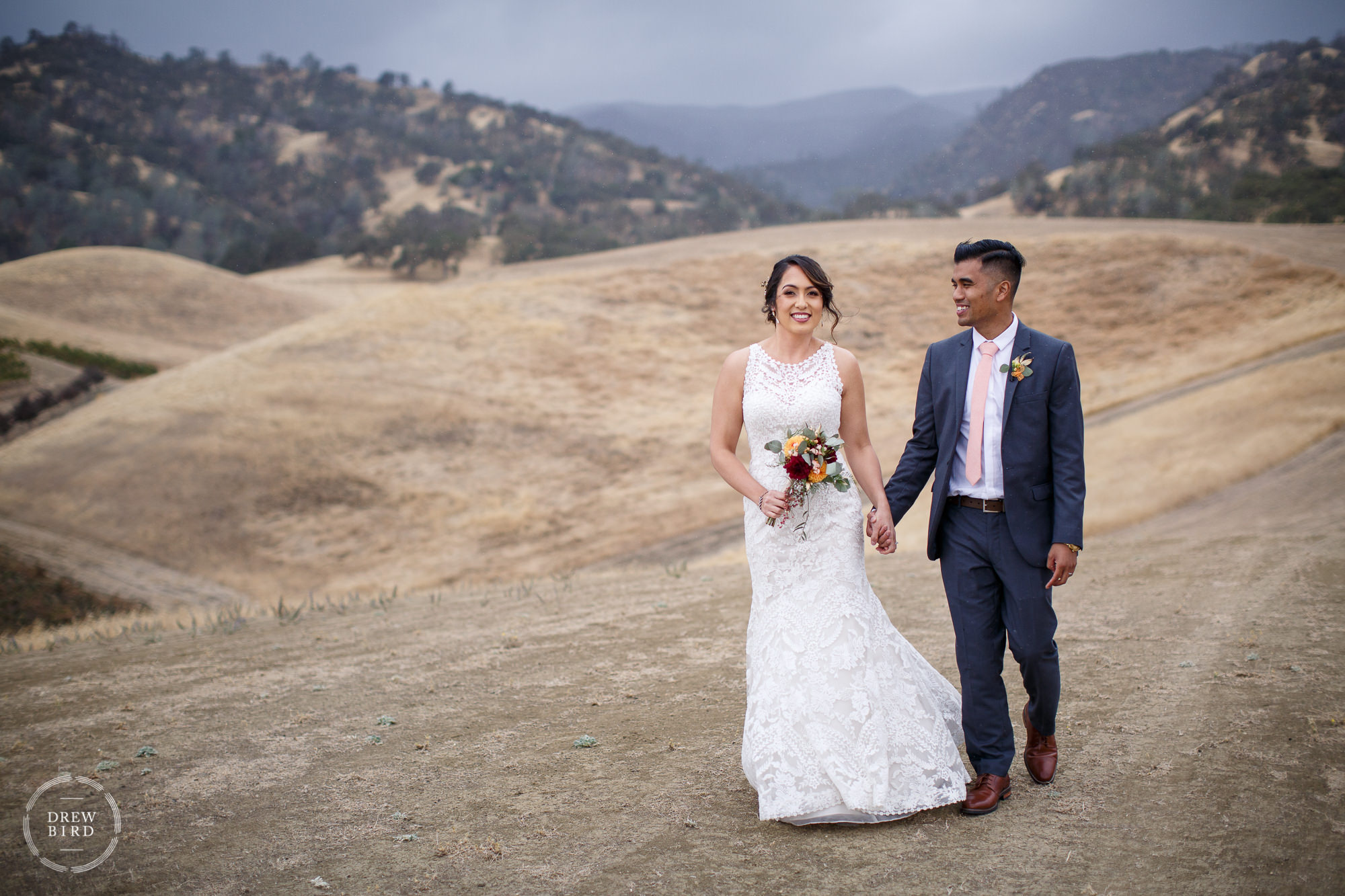 Bride and groom holding hands walking in rolling hills meadow. Taber Ranch wedding photography in Capay Valley, California.