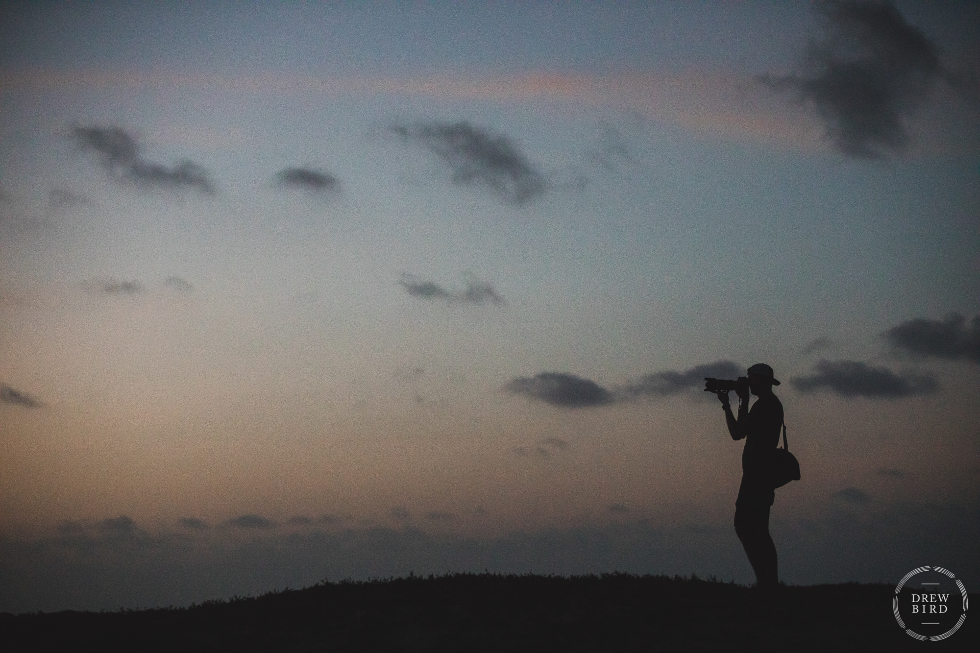 A silhouette of a photographer with camera in hand standing on a sand dune on the Caribbean island of Aruba at sunset. Photo by Destination wedding photographer Drew Bird