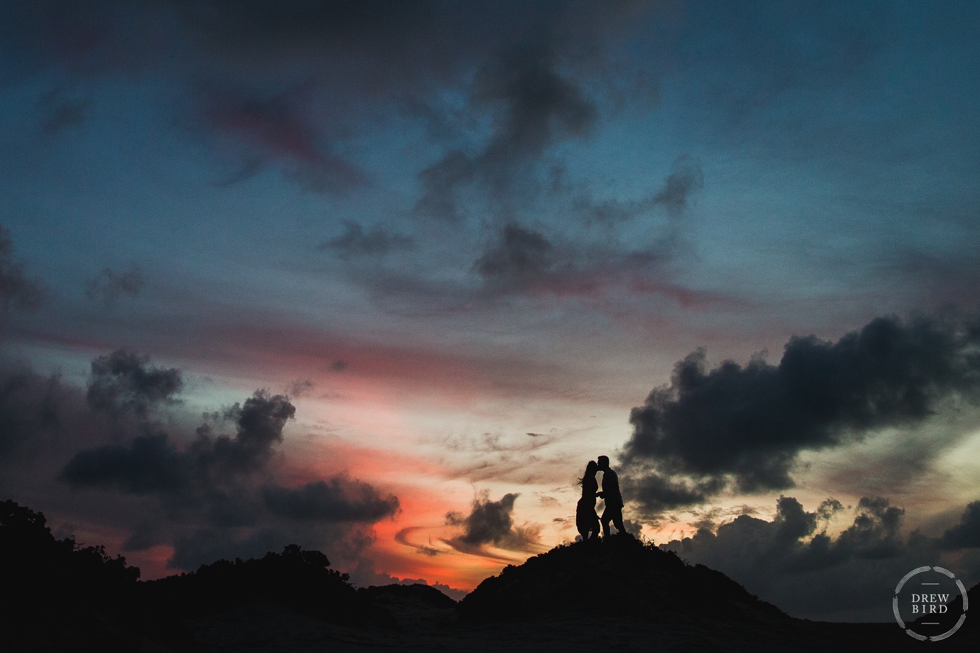 At sunset the silhouette of an Indian wedding couple atop a sand dune with mesmerizing cloud formations and gorgeous sunset colors on the tropical island of Aruba.
