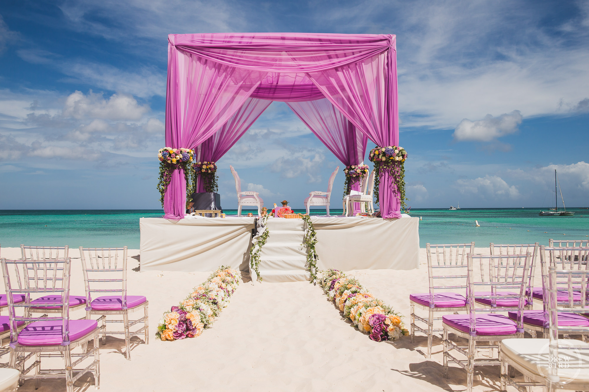 A detail photo of the stage, pink mandap, floral arrangements, and wedding seats with matching pink covers on the sand with the turquoise ocean and blue skies in the background for a Hindu wedding ceremony on the beach at the Hilton Aruba Resort in the Caribbean by Indian wedding phpotographer Drew Bird.