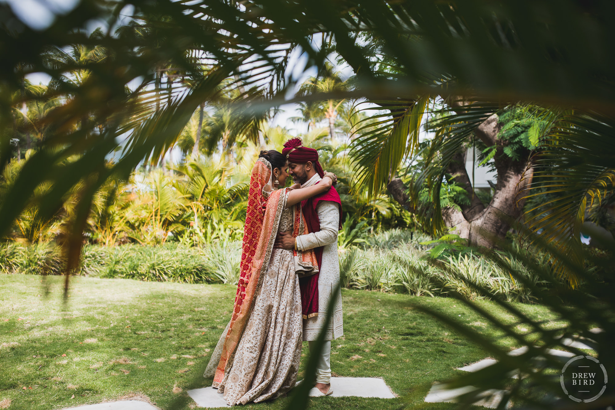 Indian wedding bride and groom embrace for the first look session at the Hilton Aruba resort in the Caribbean. The photo is creatively framed with blurred out palm leaves in the foreground for a Hindu destination wedding photography story by SF photographer Drew Bird.