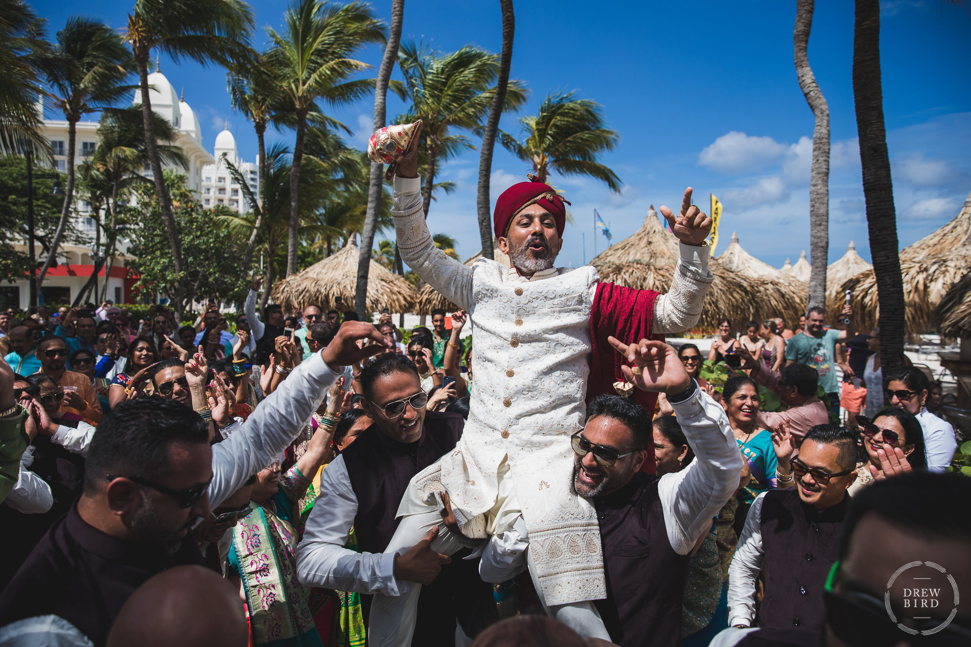 An Indian groom in full Hindu wedding attire is lifted and carried on the shoulders of his friends in a crowded celebratory parade. This is part of the baraat ceremony as the groom and his friends travel to the Indian wedding ceremony site on the beach at the Hilton Aruba resort in the Caribbean by SF Photographer Drew Bird. This is a gorgeous documentary style wedding photo of an Indian wedding ceremony.