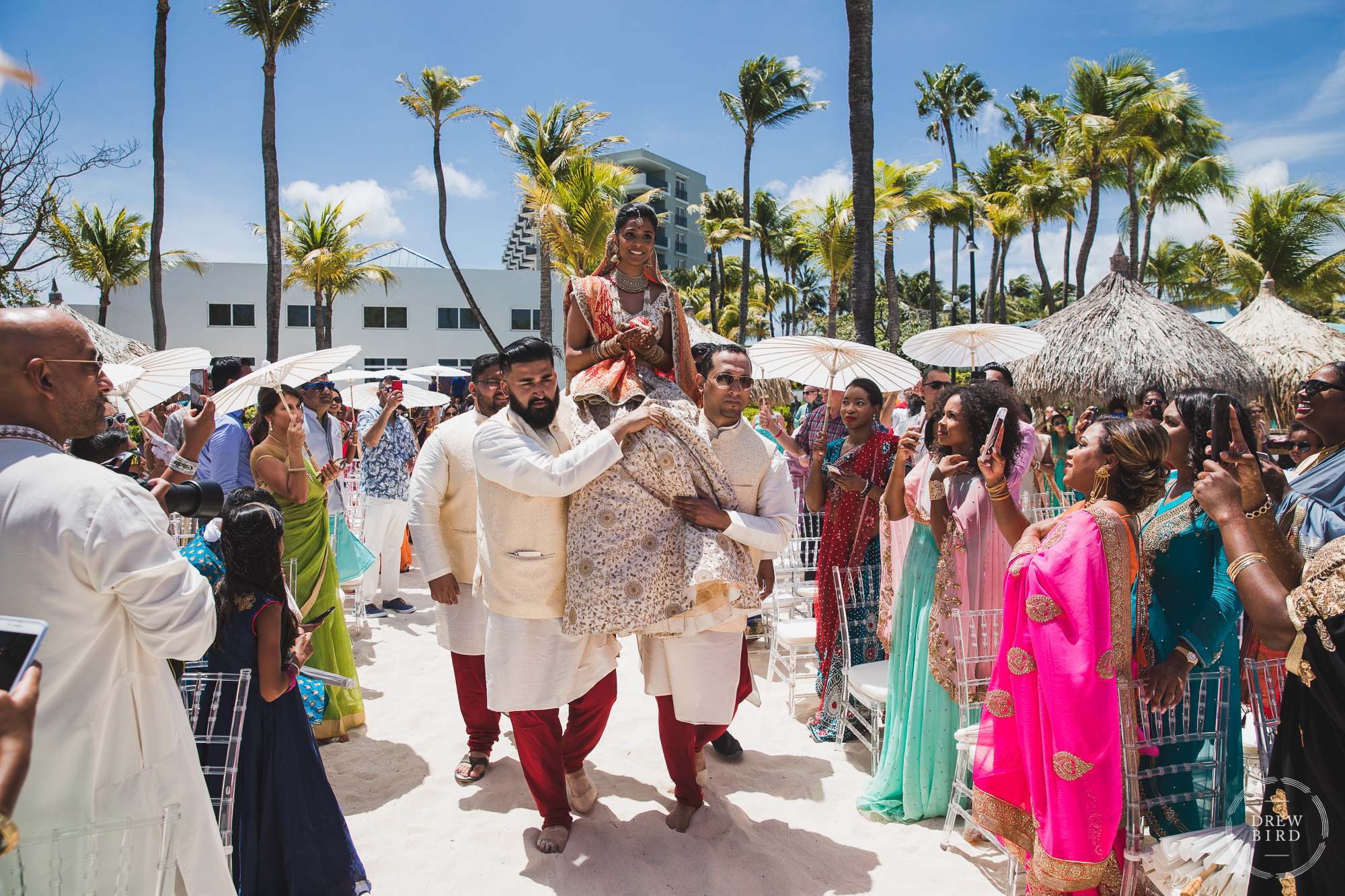 An Indian bride in her traditional Hindu wedding dress is carried on the shoulders of her family to the Mandap at the beginning of the wedding ceremony on the beach at the tropical Hilton Aruba resort in the Caribbean by Indian wedding photographer, Drew Bird.