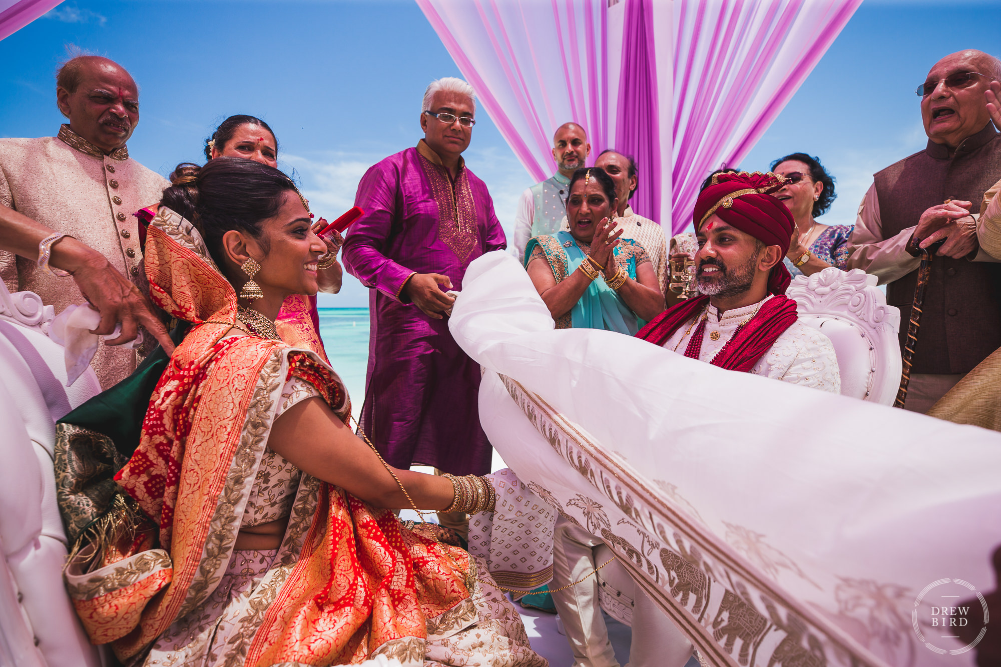 During a Hindu wedding ceremony on the beach, the bride and groom see each other for the first time as the antarpat - also called the varmala or jaimala, which is a white sheet that prevents the couple from seeing each other, is raised. The couple are holding hands and their family are standing in the background crying, laughing, and clapping. The white sheet symbolizes the two families being separate, lifting the sheet symbolizes the families being joined together. A destination Indian wedding photography story at the Hilton Aruba resort in the Caribbean by SF photographer Drew Bird.