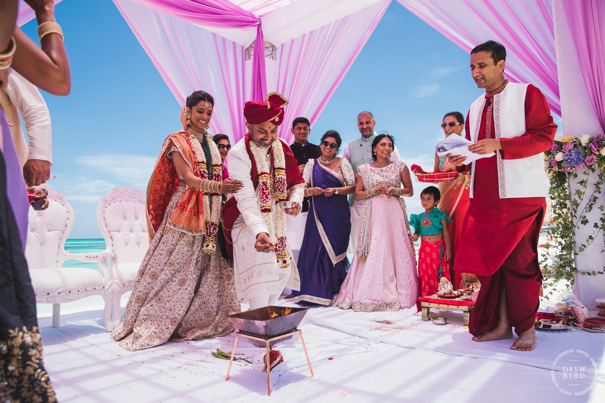 Bride and groom toss rice into an open fire under a pink Mandap with blue sky and ocean in the background for a Hindu Indian wedding ceremony on the beach at the Hilton Aruba resort by professional wedding photographer Drew Bird.