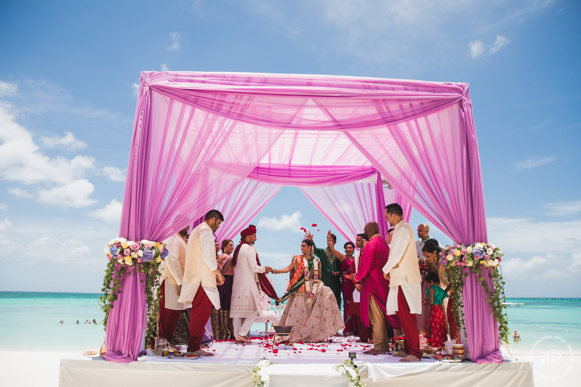 Bride and groom walk under a pink Mandap around a ceremonial fire with blue sky and ocean in the background for a Hindu Indian wedding ceremony on the beach at the Hilton Aruba resort by professional wedding photographer Drew Bird