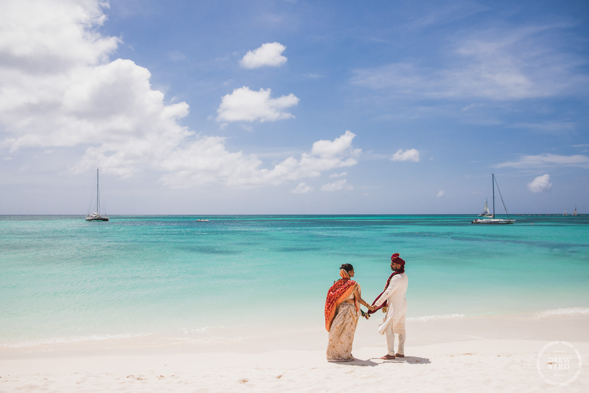 The Indian bride and groom stand on the beach at the water's edge holding hands for an artistic wedding portrait after their Hindu wedding ceremony on the beach at the Hilton Aruba Resort by SF photographer Drew Bird who specializes in wedding photography for Indian weddings.