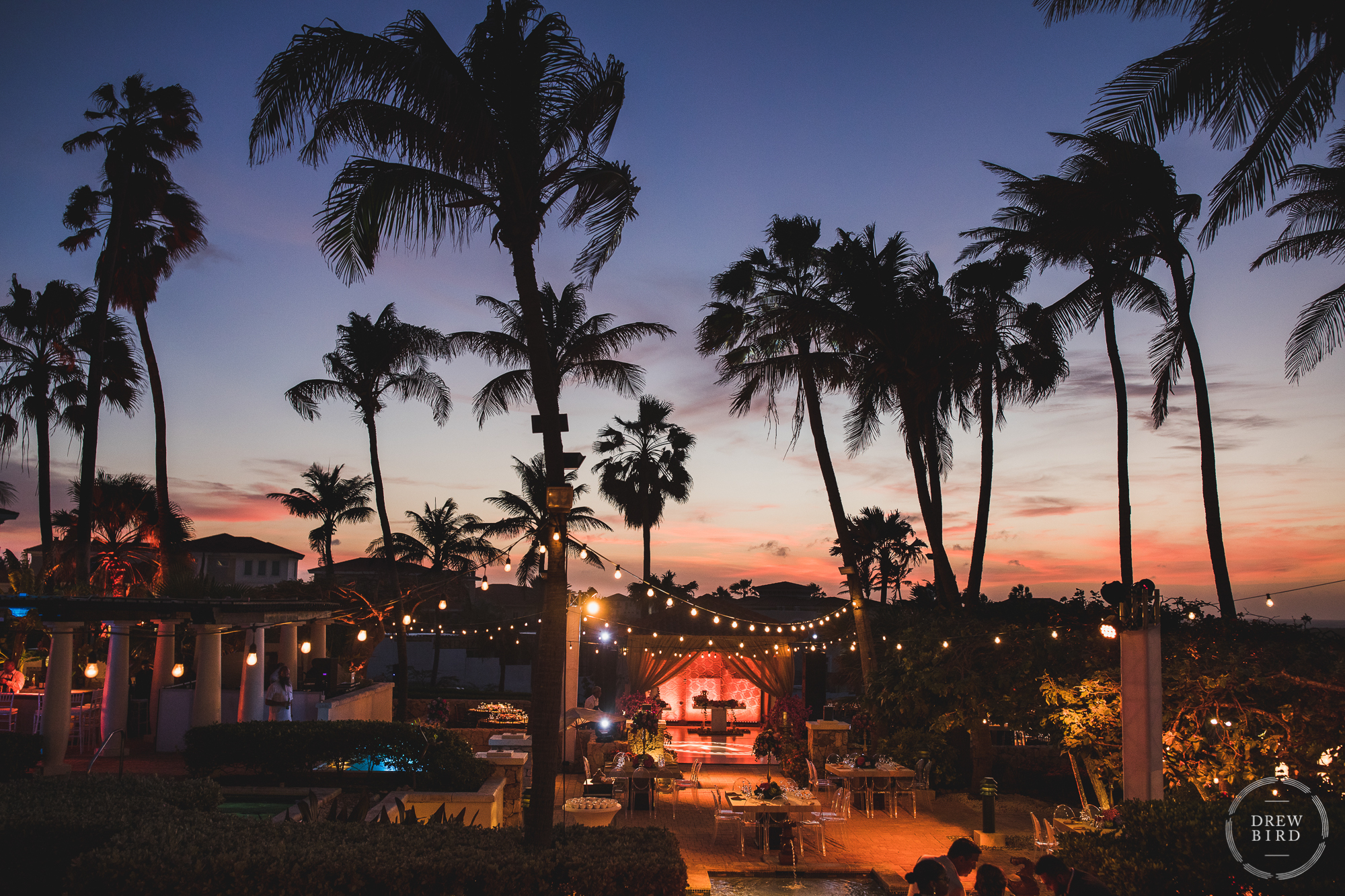 A stunning photo of Tierra del Sol, an elegant outdoor wedding reception venue and resort on the rugged north shore of Aruba in the Caribbean. Here, the dance floor and table settings are towered over by the silhouettes of palm trees against the sunset sky with colors of orange and red and pink.