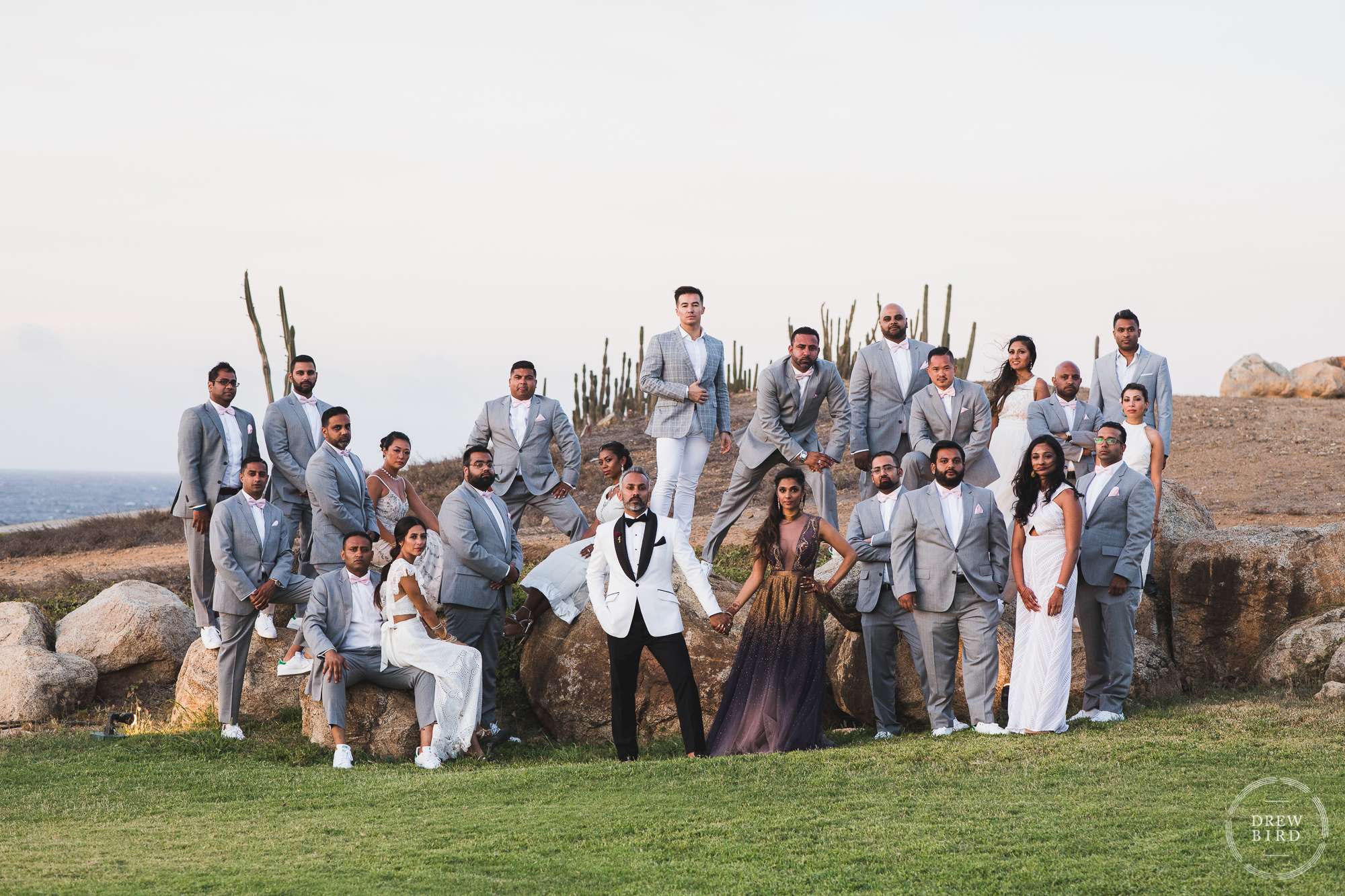 A classic and gorgeous wedding party portrait with over twenty bridesmaids and groomsmen and the Indian bride and groom. The wedding party are beautifully arranged sitting and standing on giant rock boulders at sunset at Tierra del Sol on the north shore of Aruba in the Caribbean by Aruba destination wedding photographer Drew Bird.