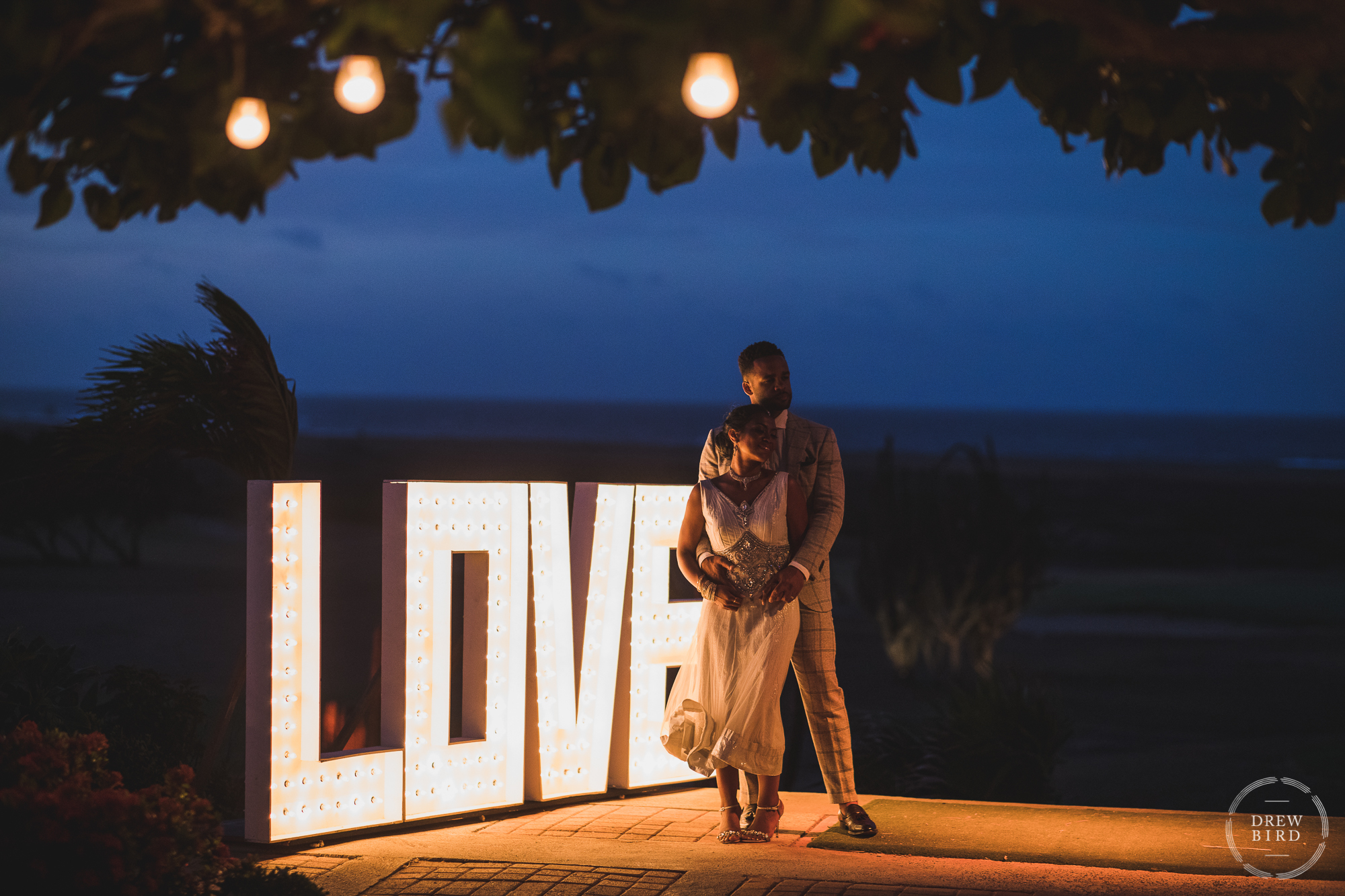 A woman stands leaning back into a man, his arms are around the woman at dusk with a large illuminated sign that says love glows in the background. The ocean is visible on the horizon. A wedding reception at Tierra del Sol in Aruba.
