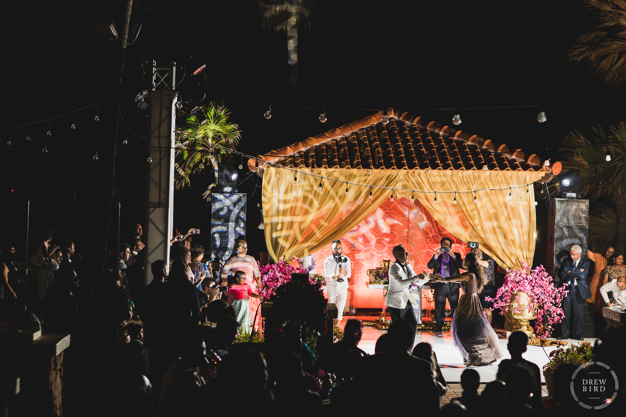 An incredible night time photo of the indian bride and groom during their first dance on the outdoor patio at Tierra del Sol, an elegant wedding venue on Aruba in the Caribbean. This photo shows the entire dance floor, buildings, and the wedding guests watching and cheering.