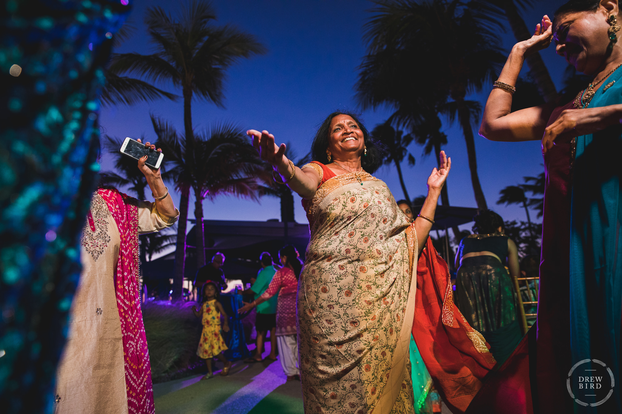 A group of women in traditional Indian wedding attire dance together at sunset. A professional wedding photography story about a Hindu destination wedding at the Hilton Aruba Resort by wedding photographer Drew Bird.