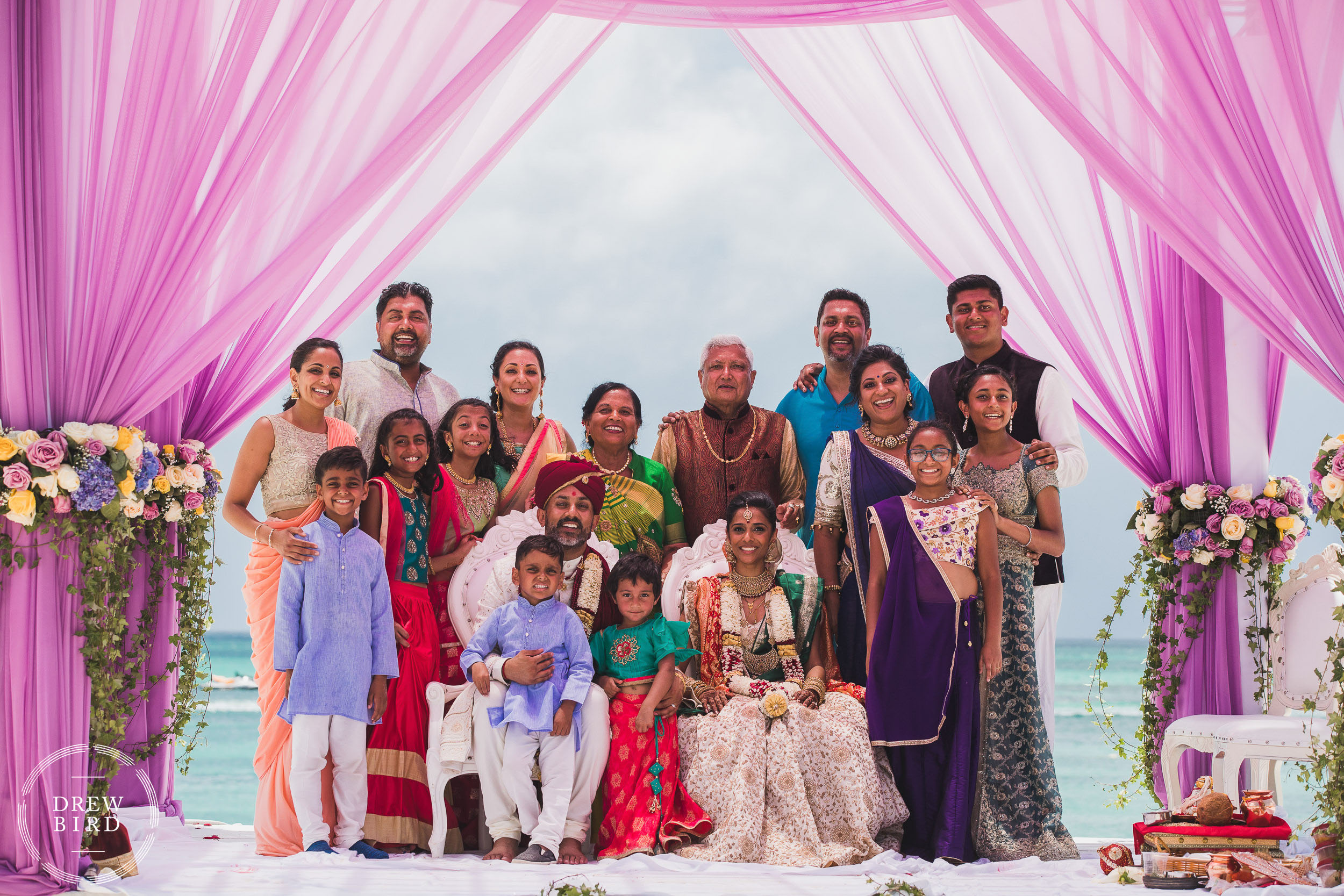 A group family portrait of a Hindu wedding couple and their family under a pink mandap on the beach with the ocean in the background following an Indian wedding ceremony at the Hilton Aruba resort in the Caribbean by SF photographer Drew Bird who specializes in documentary style Indian wedding photography.
